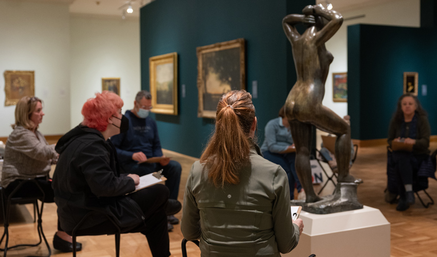 photo of seated adults in a gallery surrounding a bronze sculpture of a figure while sketching it