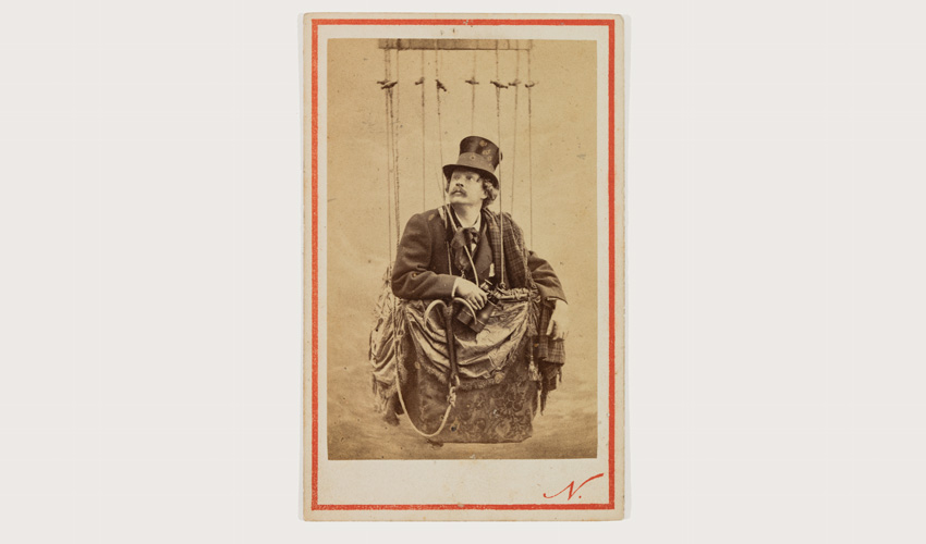 vertical portrait on card surrounded by a printed red frame of the artist in a basket hanging from an unseen balloon