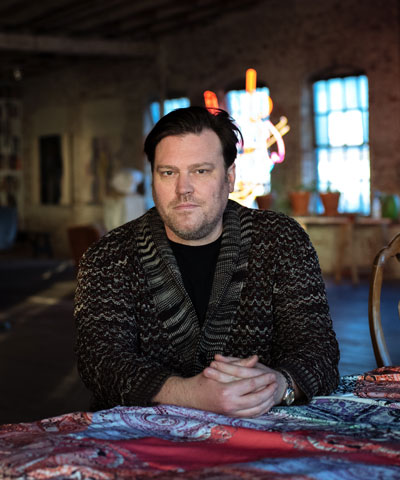photo of the artist in a dark sweater from the chest up staring into the camera with hands folded in front
