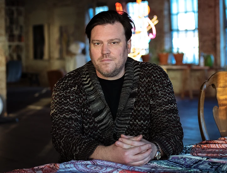 photo of the artist in a dark sweater from the chest up staring into the camera with hands folded in front