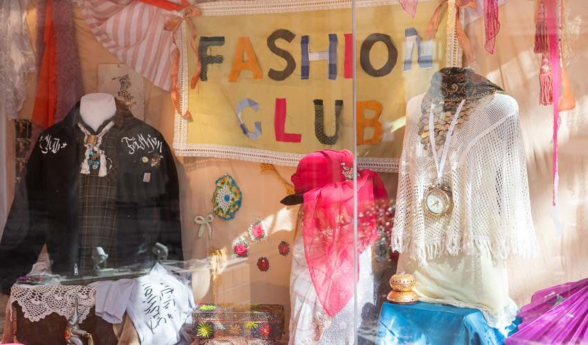 photo of display window featuring a banner with the text Fashion Club, and student designs of a black jacket, and a white top with a clock on a necklace