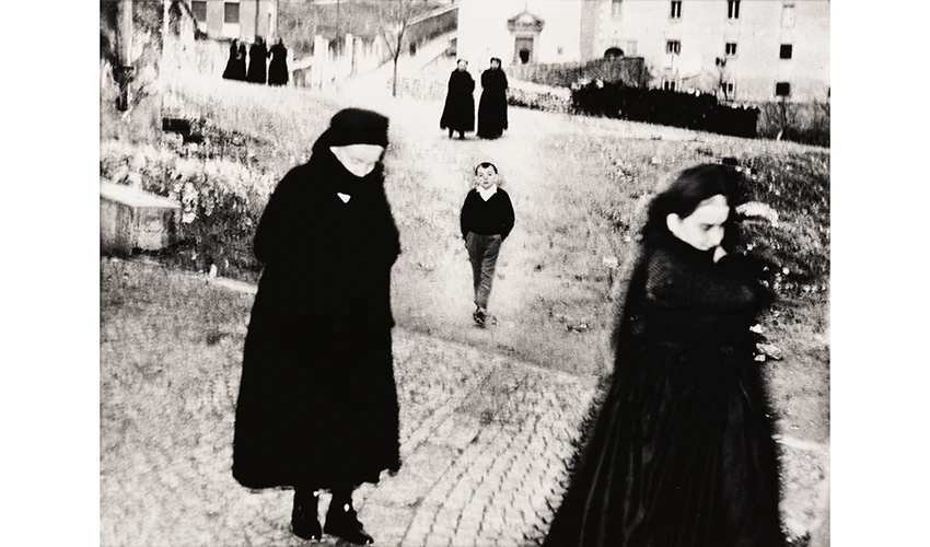 black and white photo of a street scene with black clad figures passing in the foreground framing a child walking towards the camera