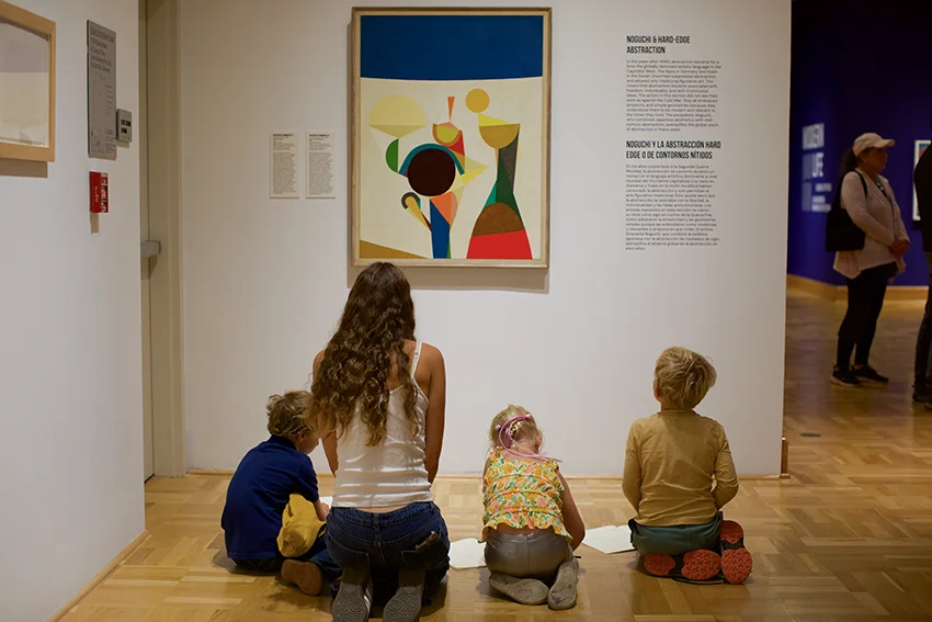 photo of a teacher kneeling surrounded by children in front of an artwork 