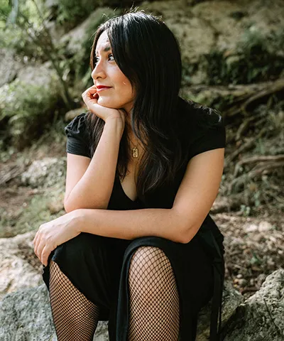 photo of presumably a woman with dark hair and a black dress siting on rocks looking to her right