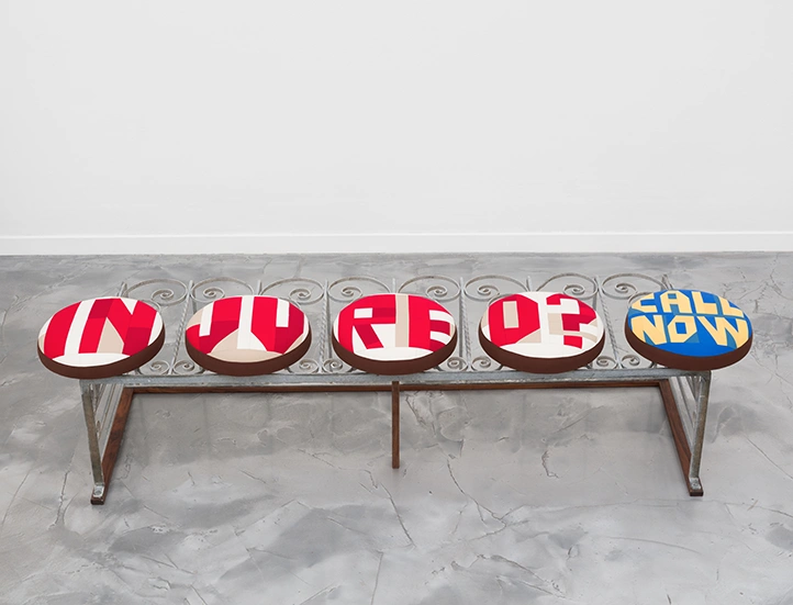 photo of a bench on a grey floor against a white wall with five round padded seats and no back with the following text on the seat covers, "injured? call now"