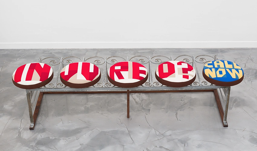 photo of a bench on a grey floor against a white wall with five round padded seats and no back with the following text on the seat covers, "injured? call now"