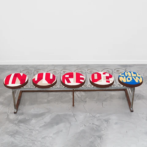 photo of a bench on a grey floor against a white wall with five round padded seats and no back with the following text on the seat covers, "injured? call now"