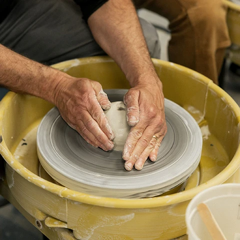 photo of a pair of lighter colored hands working clay in a yellow pottery wheel