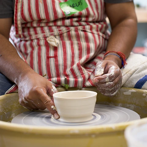 photo of a darker colored person in a red and white striped apron at a pottery wheel working clay