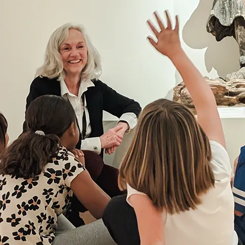photo of older docent woman smiling in front of a group of children while one raises their hand