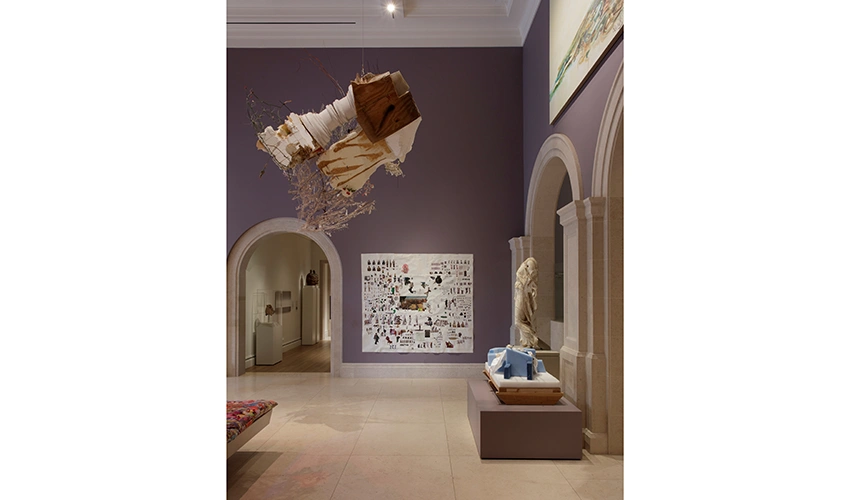 Image of art gallery with purple walls. Sculpture hanging from ceiling. Ancient Rome marble statue partially left in his packing material. Collage by artist Elliott Hundley with cut out of sculptures from the Santa Barbara Museum of Art collection