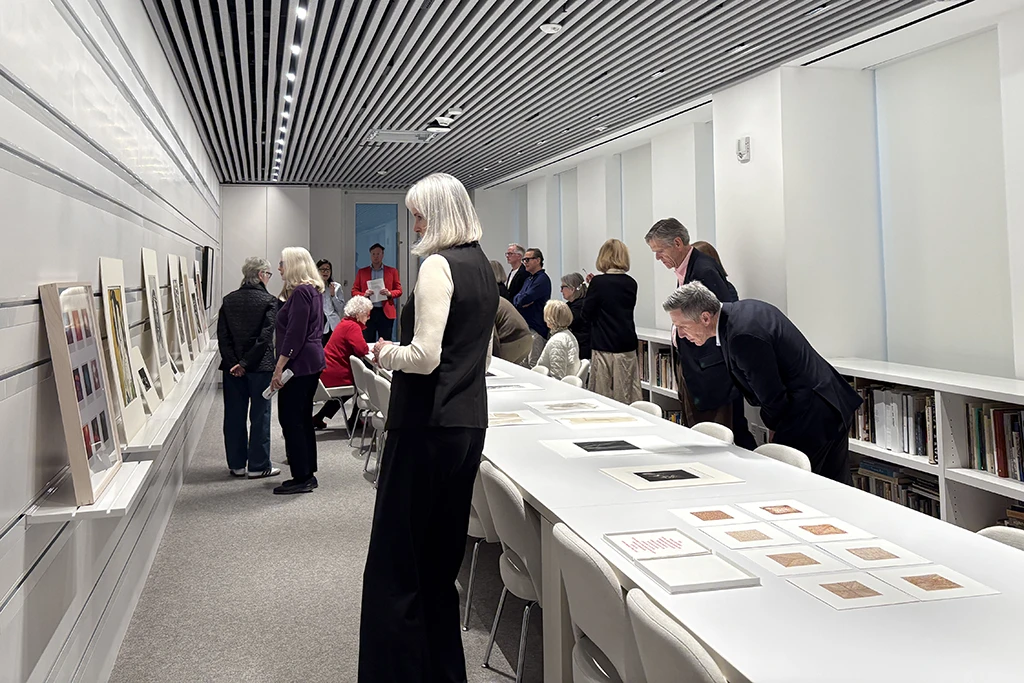 people in a modern interior of light gray walls looking at art on the walls and long table that runs through the frame