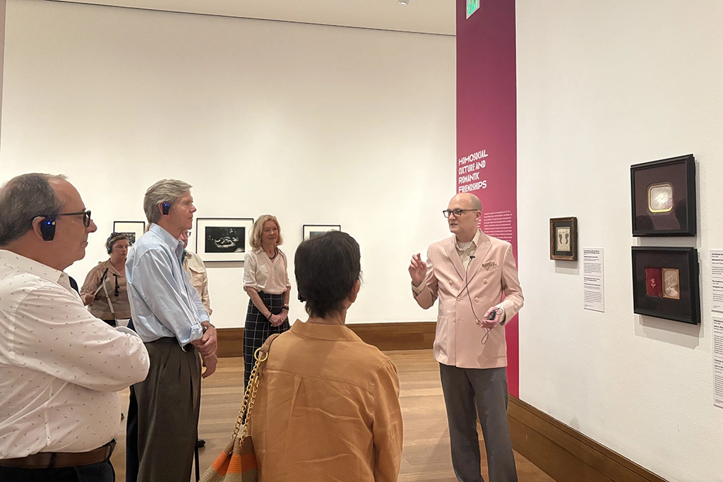 group of well dressed museum visitors in a gallery getting a tour from a balding man in a pink sport coat
