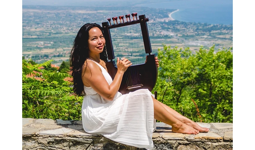 photo of, presumably, a woman with dark hair in a white dress sitting on a stone wall holding up a lyre with a coastal landscape behind them
