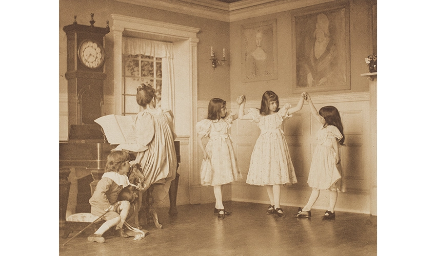 monochrome photo of three girls in dresses dancing along to a woman at a piano and a little boy with a string instrument looking on