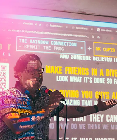 photo of a man in front of a screen being projected with lyrics and graphics speaking into a microphone