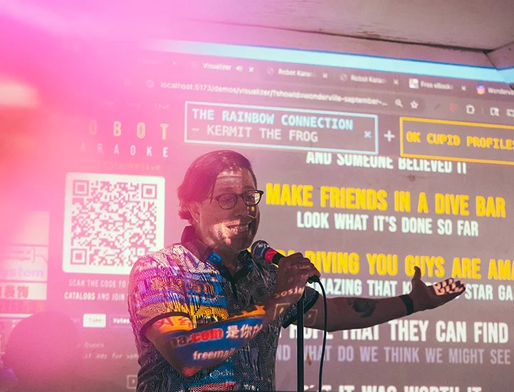 photo of a man in front of a screen being projected with lyrics and graphics speaking into a microphone