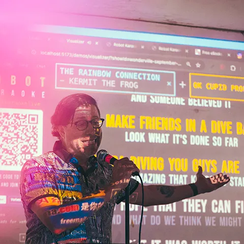 photo of a man in front of a screen being projected with lyrics and graphics speaking into a microphone