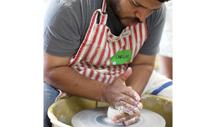 a bearded man wearing an apron working on ceramics at a potters wheel 