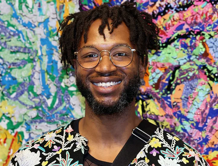 Black man with glasses, short hair, and a beard, wearing a colorful floral print jacket, smiling into the camera photographed against a colorful and abstract backdrop