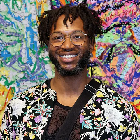Black man with glasses, short hair, and a beard, wearing a colorful floral print jacket, smiling into the camera photographed against a colorful and abstract backdrop