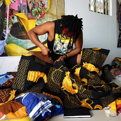 photo of a seated Black man in a dark cutoff tee cutting ornate fabric