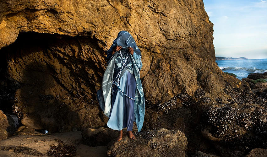 photo of a dark skinned person wrapped in indigo colored fabric standing against a rocky seaside 