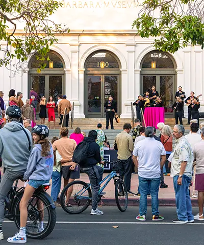 A crowd of visitors, including cyclists, gathers on the sidewalk in front of the Santa Barbara Museum of Art at dusk.