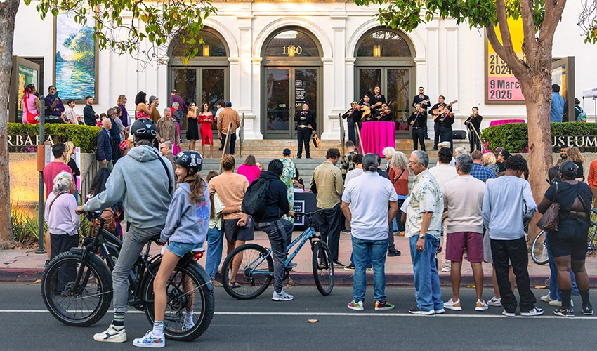A crowd of visitors, including cyclists, gathers on the sidewalk in front of the Santa Barbara Museum of Art at dusk.