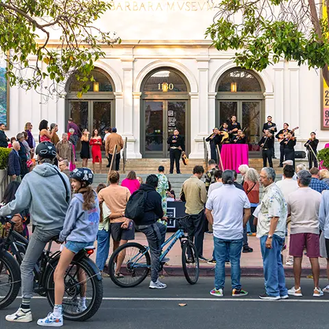 A crowd of visitors, including cyclists, gathers on the sidewalk in front of the Santa Barbara Museum of Art at dusk.