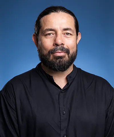 photo of a dark haired bearded man in a dark collared shirt against a blue background