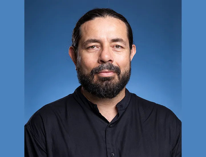 photo of a dark haired bearded man in a dark collared shirt against a blue background