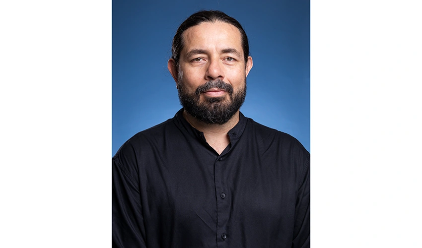 photo of a dark haired bearded man in a dark collared shirt against a blue background