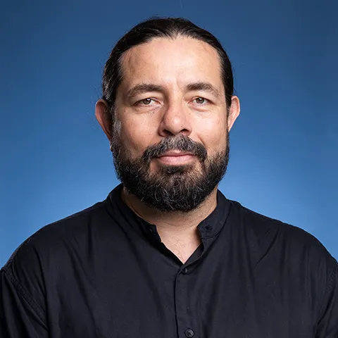 photo of a dark haired bearded man in a dark collared shirt against a blue background