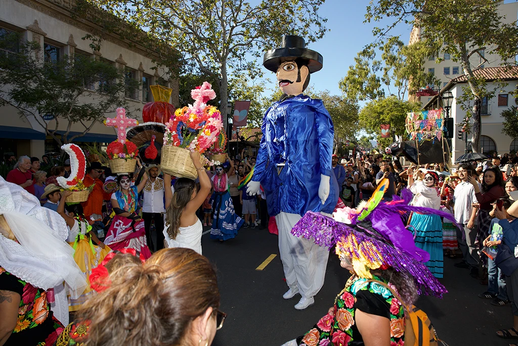 two giant puppets surrounded by a crowd of people on the street in front of the museum on a sunny day