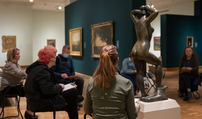 photo of seated adults in a gallery surrounding a bronze sculpture of a figure while sketching it
