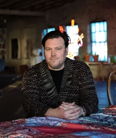 photo of the artist in a dark sweater from the chest up staring into the camera with hands folded in front