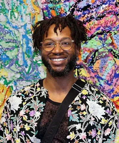 Black man with glasses, short hair, and a beard, wearing a colorful floral print jacket, smiling into the camera photographed against a colorful and abstract backdrop