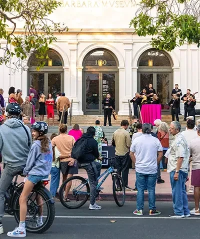 A crowd of visitors, including cyclists, gathers on the sidewalk in front of the Santa Barbara Museum of Art at dusk.