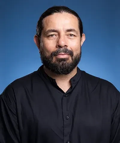 photo of a dark haired bearded man in a dark collared shirt against a blue background