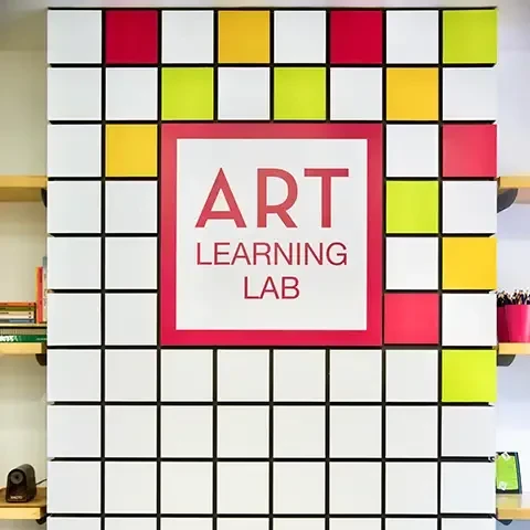 photo the Art Learning Lab sign and display featuring white checkerboard partially filled in with red, orange, and green flanked by shelves filled with supplies