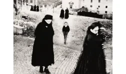 black and white photo of a street scene with black clad figures passing in the foreground framing a child walking towards the camera