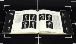 video still of a black surface with white registration marks and a book of black and white photos of a performer laid open on top