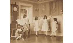 monochrome photo of three girls in dresses dancing along to a woman at a piano and a little boy with a string instrument looking on